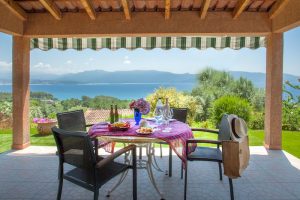 Location corse 2023 - Résidence de vacances pas chère Cantu Di Mare. Image de la terrasse de la résidence de vacances avec, en premier plan, une table sur laquelle nous pouvons voir un vase rempli de fleurs et deux bouteilles de champagnes et des chaises, donnant vue sur les montagnes corses. 
